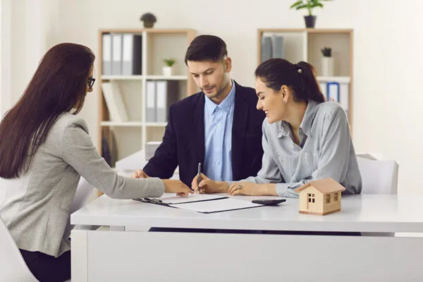 Three adults review paperwork at a desk while one woman signs a document beside a wooden