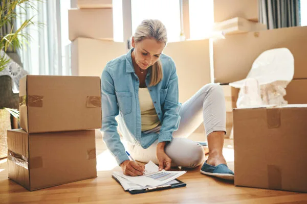 Woman writing on paperwork attached to a clipboard while sitting beside cardboard boxes.