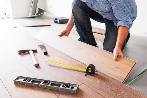 A person installing hardwood flooring, with tools such as a hammer, tape measure, and level placed nearby on the floor.