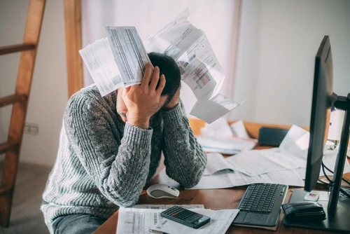 A frustrated man sitting at a desk, holding his head, surrounded by scattered bills and papers.