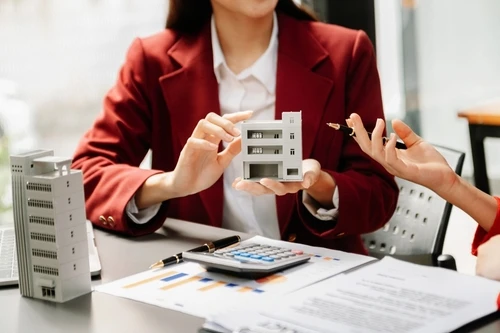 Two professionals are sitting at a desk, discussing a miniature model of a building. One person holds the model, while the other gestures with a pen. Documents, charts, and a calculator are spread out on the table, indicating a conversation about real estate or property management.