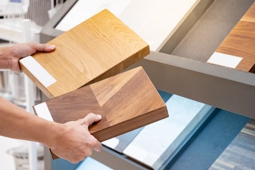 A person holding two wooden flooring samples while selecting from a display of various flooring options.