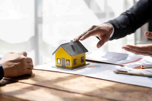 Two individuals are seated at a table, discussing a small model of a yellow house placed on top of documents and charts. One person is gesturing toward the model, suggesting a conversation about real estate, property investment, or home purchasing.