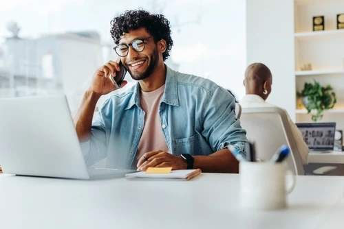 A smiling man sitting at a desk, talking on the phone while using a laptop, in a bright office setting.