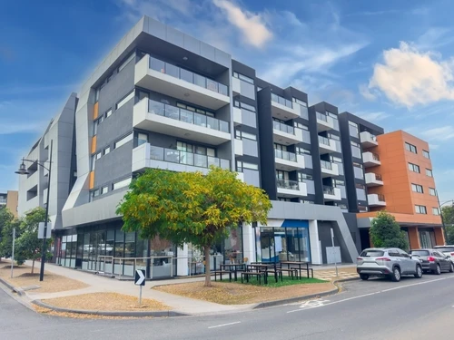 A modern multi-story apartment building with large balconies and a ground-floor commercial space is situated on a street corner. Several cars are parked nearby, and a tree is visible in front of the building under a clear sky.