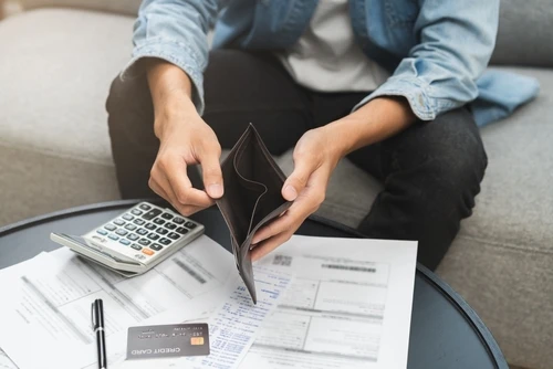 A person sitting at a table, holding an empty wallet with bills, a calculator, and a credit card on the table in front of them.