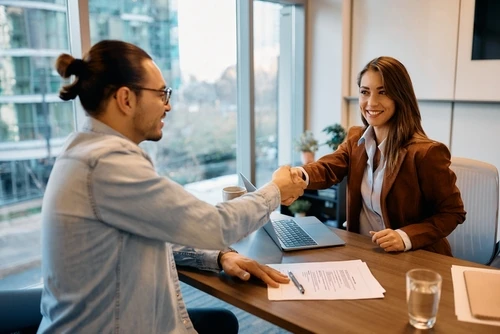 A man and a woman smiling and shaking hands across a desk, with documents and a laptop in front of them.