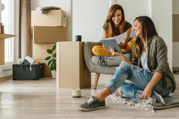Two women sit in an apartment with moving boxes talking about taking over a lease.