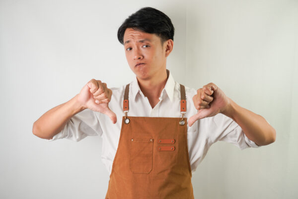 Young man wearing rolled-up white sleeve shirt and brown apron with thumbs down hand gesture. 