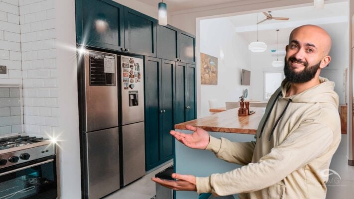 Smiling man presenting a modern kitchen with stainless steel appliances, teal cabinets, & a wooden dining area.