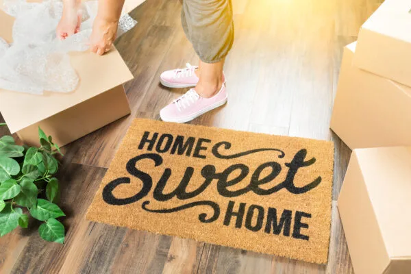 Woman in Pink Shoes and Sweats Unpacking Near Home Sweet Home Welcome Mat, some Moving Boxes, and a Plant.