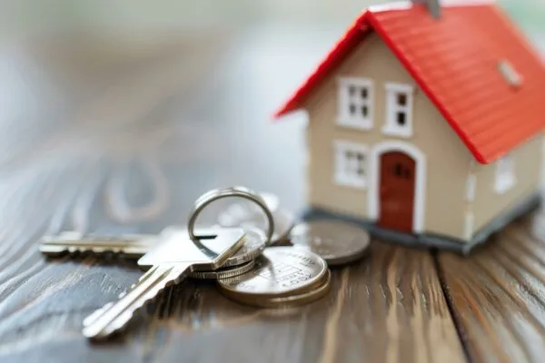 House keys are resting on a stack of coins with a miniature house out of focus in the background