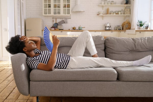 Tired woman suffering from heatstroke flat without air-conditioner, waving blue fan, lying on couch at home. Black girl cooling in hot summer weather. Overheating, high temperature.