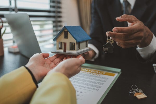 A landlord handing over a model house and keys to a tenant across a desk with a rental contract in view