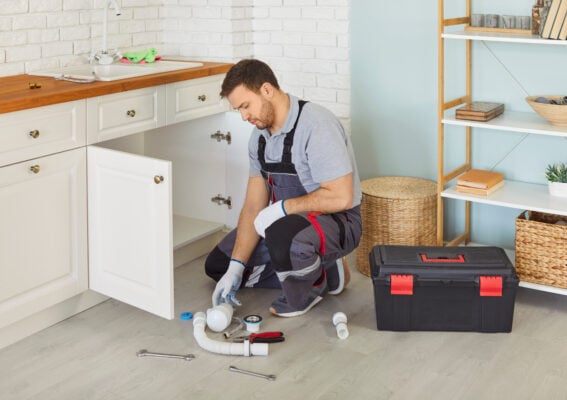 Professional male plumber in uniform with toolbox sitting near sink replacing the water filter or fixing broken pipe in modern kitchen. Repair service and plumbing maintenance concept.