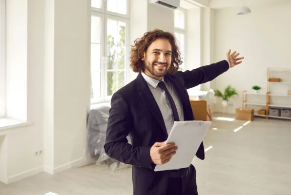 POV point of view shot of happy friendly man professional real estate agent in suit with clipboard giving tour about new modern house or apartment, doing welcoming gesture, showing customers interior