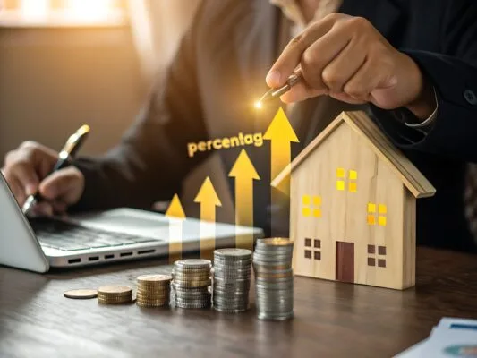 Stacks of coins and a wooden house model with rising arrow graphics, symbolizing increasing ROI in real estate investment.