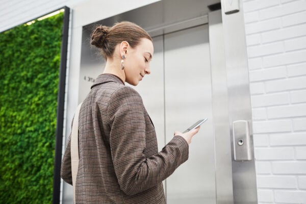 Minimal low angle shot of young woman waiting for elevator in modern building and using phone, copy space
