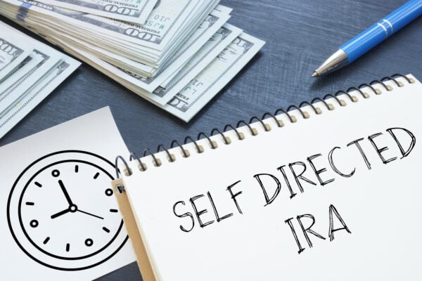 Notebook with the words Self Directed IRA surrounded by money, clock, and pen, symbolizing retirement investment options