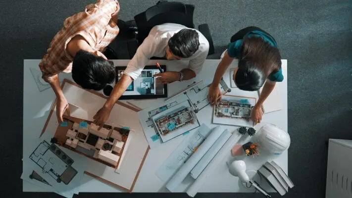 Top view of project manager working together to brainstorm house design while standing around meeting table with project plan and blueprint. Aerial view of designer team explain project.