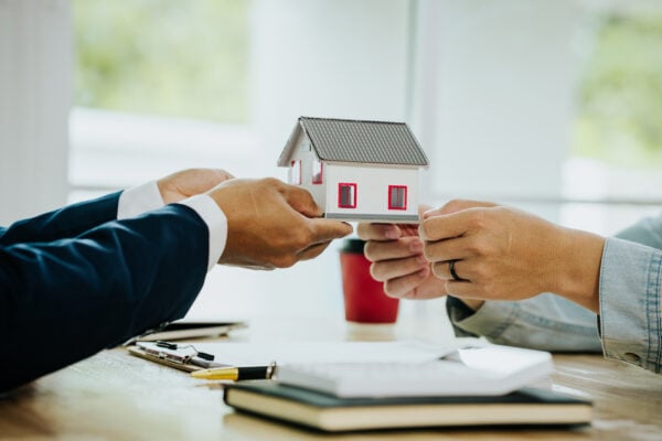 Two people exchanging a small house model across a table, symbolizing a real estate contract transfer or property assignment.