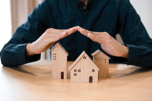 Hands covering small wooden houses symbolizing blanket insurance coverage for multiple properties.