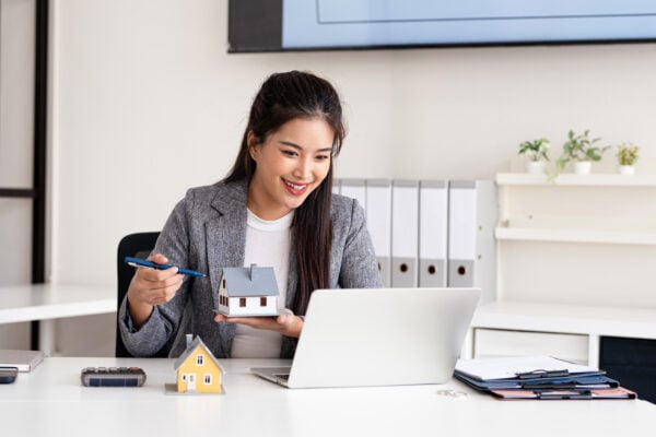 A real estate professional holding a model house while working on a laptop, representing guidance in choosing different types of property deeds.