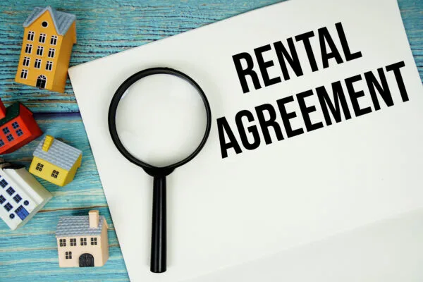 Notepad with "Rental Agreement" with magnifying glass and house model on wooden background