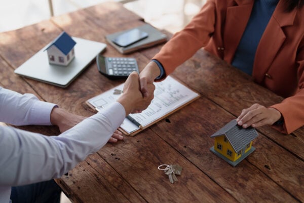 landlord and property manager seal a property deal with a handshake over a wooden desk strewn with house models, keys, calculator and contract, symbolizing real estate transaction success