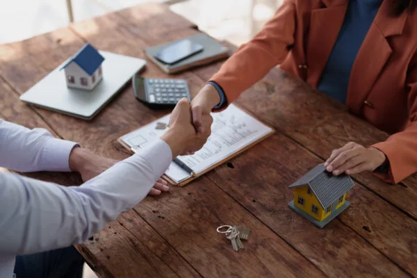 landlord and property manager seal a property deal with a handshake over a wooden desk strewn with house models, keys, calculator and contract, symbolizing real estate transaction success